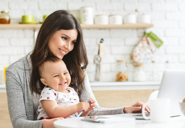 Jovem mãe digitando no laptop, sentado com o bebê na cozinha
 - Foto, Imagem