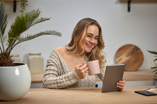 Model blogger in glasses, beige sweater. Smiling, holding tablet, pink cup. Sitting in kitchen at table with palm, smartphone, plastic card. Close-up - Photo, Image