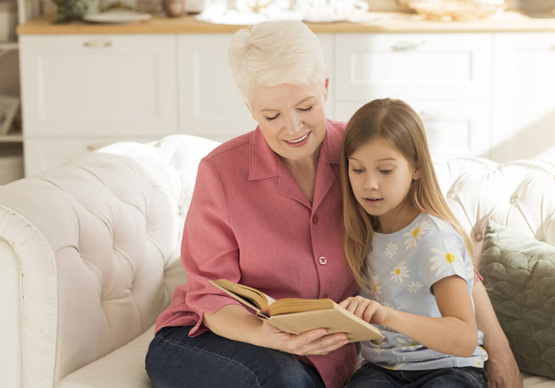Elderly lady and her granddaughter with interesting book on sofa at home - Foto, immagini