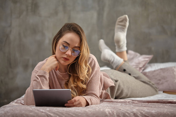 Blondynka w okularach, luźne ubranie. Leżąc na łóżku, podpierając jej głowę ręką, pracując z tabletem. Studentka, blogerka. Zbliżenie - Zdjęcie, obraz
