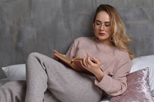 Blonde female in glasses, casual clothing is reclining on bed, reading book in bedroom. Student, blogger. Interior with gray wall. Close-up - Foto, Imagem