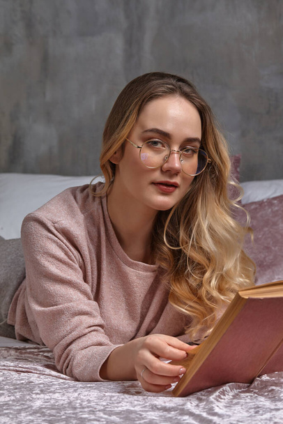 Blonde lady in glasses, casual clothing. She is looking at you, holding book, laying on bed in bedroom. Student, blogger, document studying. Close-up - Zdjęcie, obraz