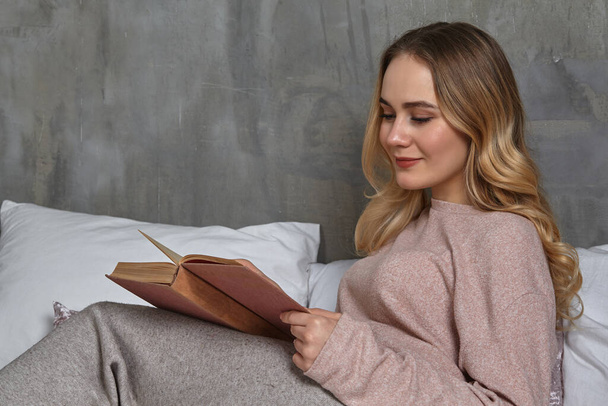 Female in casual clothing. Smiling, laying on bed, reading book in bedroom. Student, blogger, document studying. Interior with gray wall. Close-up - Zdjęcie, obraz