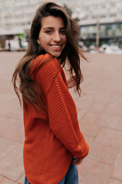 Outdoor shot of good-looking european female model posing on city background with pleasure. woman in orange pullover laughing in cold spring morning.  - 写真・画像