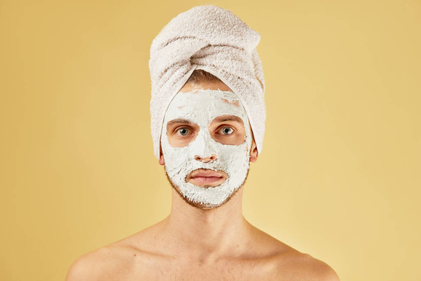 Portrait of handsome unshaven man with towel on his head and white mask on face in the studio isolated on yellow background, skin care concept - Photo, Image