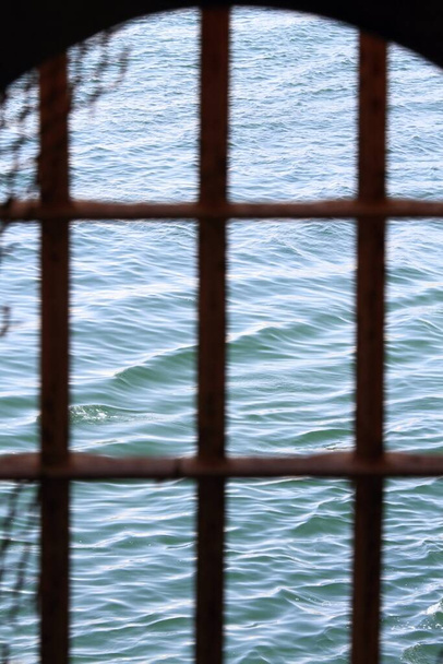 Rusty metallic bars with wire in the window. Fence bars  and rusty net, the sea behind. Quarantine does not work.  - Photo, Image
