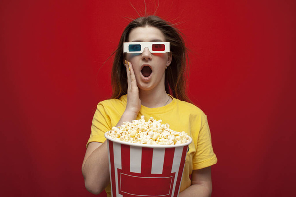 funny young girl student watching a movie in 3D glasses and screaming on a red background, she eats popcorn and is delighted, copy space - 写真・画像