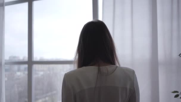 Back view of young sensual Caucasian woman coming to floor-to-ceiling window in the morning. Brunette thoughtful girl looking out indoors. Lifestyle, leisure, joy. - Πλάνα, βίντεο