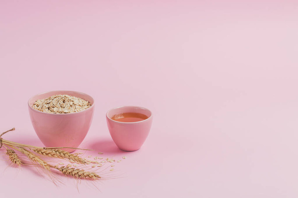 Ingredients for making homemade facial mask or scrub. Bowl of dry oat flakes with honey and ears of wheat on light background. Beauty concept. - Photo, image