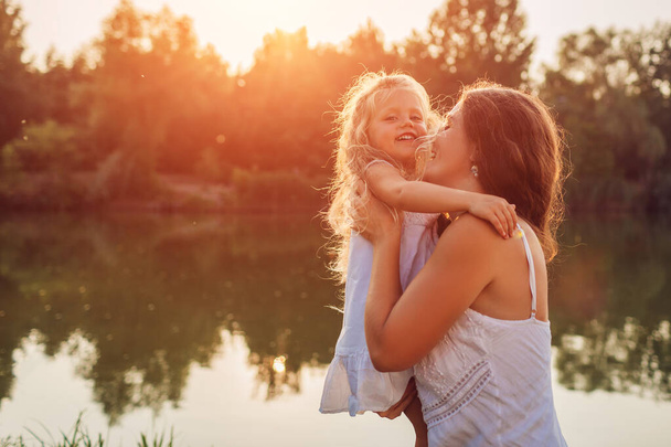 La fête des mères. Femme jouant s'amuser avec sa fille par la rivière d'été au coucher du soleil. Femme tenant un enfant et riant. Valeurs familiales
 - Photo, image