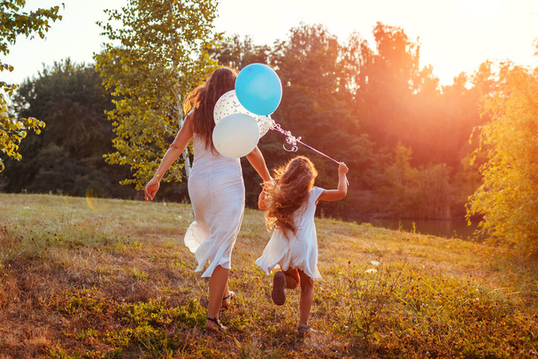母の日だ。母と一緒に走る幸せな女の子と手に風船を持っています。日没の夏の公園で家族で楽しんでいます。国際子供の日 - 写真・画像