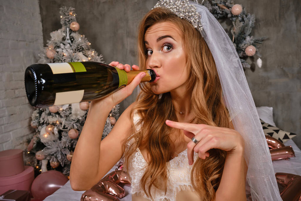 Woman in beige lingerie and white veil enjoying hen-party of bride, sitting on bed, drinking champagne. Christmas decorations, balloons. Close-up. - Zdjęcie, obraz