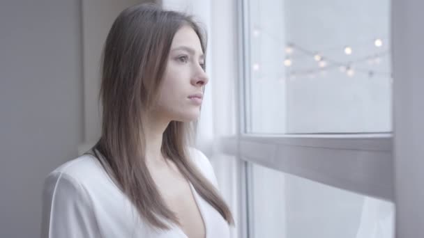 Close-up of thoughtful brunette Caucasian woman with brown eyes looking out the window at home. Cute girl waking up in the morning on weekends. Lifestyle, joy, leisure. - Séquence, vidéo