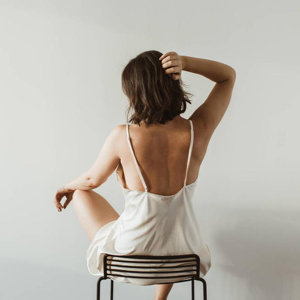 Young pretty woman in silk underwear sitting on black stool on white background. Fashion concept. - Фото, изображение