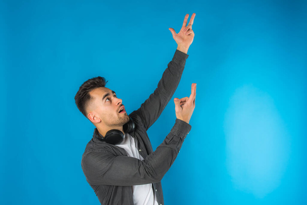 portrait of handsome european young man in gray shirt and white t-shirt with big black headphones on blue background, emotional, showing up with hands and looking - Photo, Image