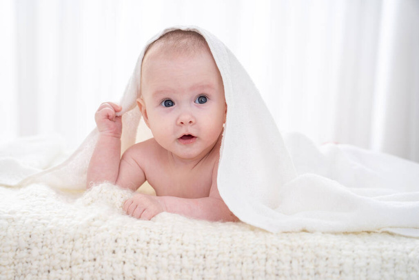 Sweet baby is lying on the bed under a white blanket. small curious child 4 months, Caucasian, lies on the surface in bright cozy room, covered with light blanket and looks away on a white background - 写真・画像