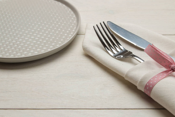 Накрываем стол. тарелка и столовые приборы в белой салфетке с розовой лентой, вилкой и ножом на белом деревянном столе
.  - Фото, изображение