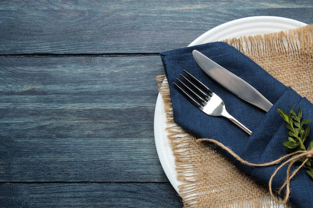 Накрываем стол. тарелка и столовые приборы в голубой салфетке, вилка и нож на голубом деревянном столе. вид сверху
 - Фото, изображение