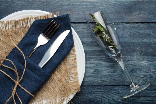 Накрываем стол. тарелка, стекло и столовые приборы в голубой салфетке, вилка и нож на голубом деревянном столе. вид сверху
 - Фото, изображение