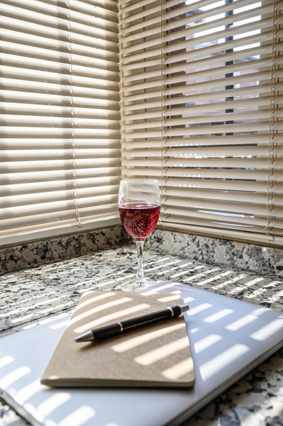 Бокал красного вина на кухне с ноутбуком, блокнотом и ручкой. Концепция отдыха дома. Вертикальное пространство копирования
 - Фото, изображение