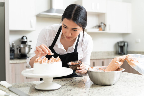 Dedicated attractive young baker sprinkling chocolate on cake in kitchen at home - Photo, Image