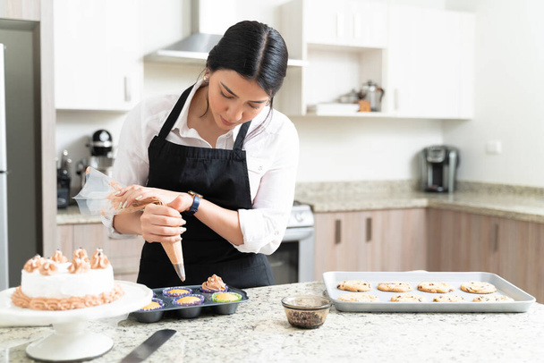 Уверенный молодой кондитер украшает кексы в кастрюле на кухонном столе дома
 - Фото, изображение