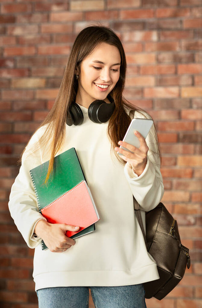 Girl with notebooks looking at smartphone and smiling - Foto, afbeelding