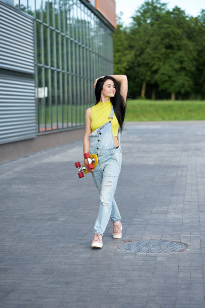 Menina morena feliz atraente com cabelos longos vestindo top amarelo e macacão jeans azul na moda andando com skate amarelo com rodas vermelhas. Conceito de estilo de vida ativo e esportivo
.  - Foto, Imagem