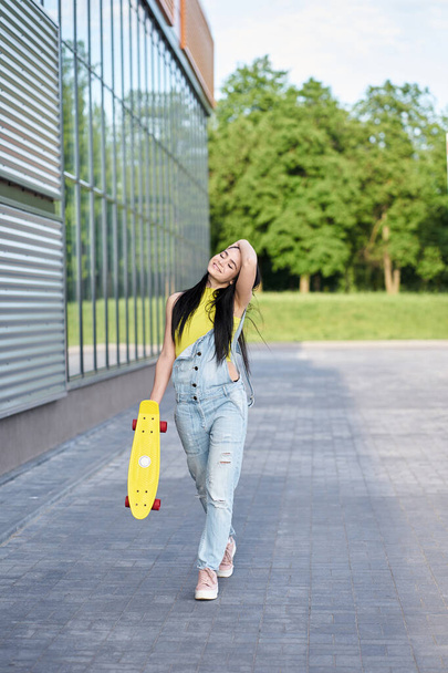 Joyful happy brunette girl with long hair wearing yellow top and modern blue denim overalls walking and holding yellow skateboard with red wheels. Active and trendy lifestyle concept.  - 写真・画像