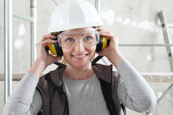mujer sonriente trabajador retrato con casco, gafas de seguridad y audífonos de protección auditiva, andamiaje interior sitio de construcción de fondo
 - Foto, imagen