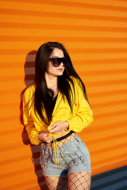 Modieuze leuke jonge vrouw draagt stijlvolle jeugd gele jas, spijkerbroek en zonnebril poseren op oranje stedelijke muur achtergrond.                                - Foto, afbeelding