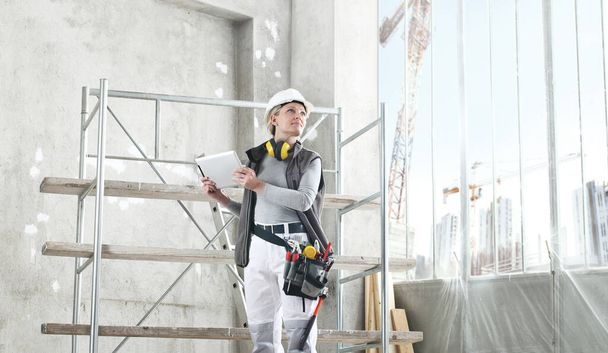 női munkás építész munka digitális táblagép, visel sisak, hallásvédő fejhallgató és táska szerszámok, állványzat építési telek beltéri háttér - Fotó, kép