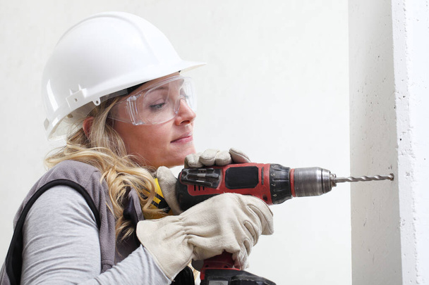 Bauarbeiterin mit Akkubohrschrauber, der ein Loch in die Wand bohrt, Bauarbeiterin mit Schutzhelm, Kopfhörer, Handschuhe und Schutzbrille, Nahaufnahme Porträt - Foto, Bild