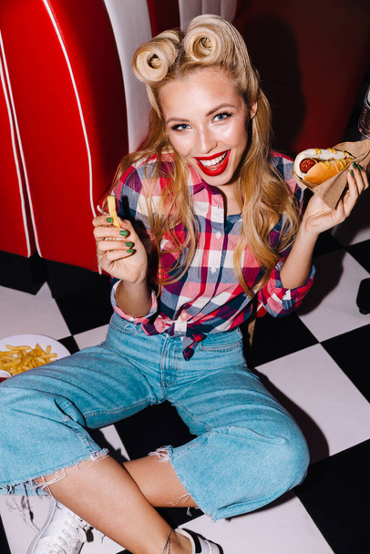 Photo of young joyful woman with beautiful hairstyle eating hotdog and french fries in retro american cafe - Foto, immagini