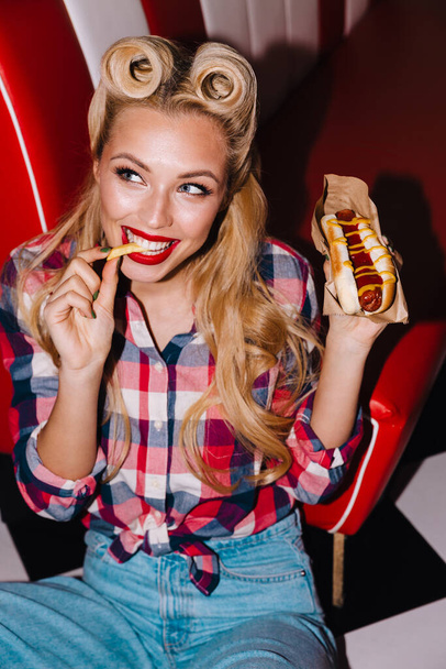 Photo of young joyful woman with beautiful hairstyle eating hotdog and french fries in retro american cafe - Φωτογραφία, εικόνα