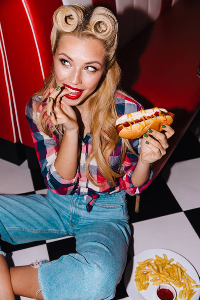 Photo of young excited woman with beautiful hairstyle eating hotdog and french fries in retro american cafe - Zdjęcie, obraz