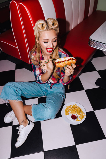 Фото молодої збудженої жінки з красивою зачіскою, що їсть хот-дог і картоплю фрі в ретро-американському кафе
 - Фото, зображення