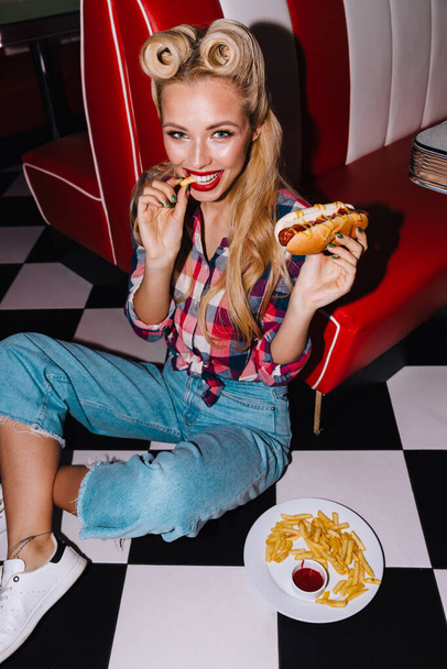Photo of young laughing woman with beautiful hairstyle eating hotdog and french fries in retro american cafe - Zdjęcie, obraz