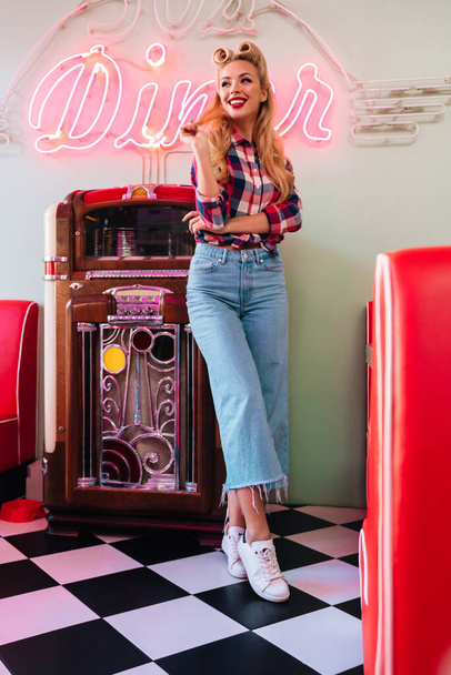 Photo of young joyful woman with beautiful hairstyle smiling while leaning jukebox in retro american cafe - Foto, Imagen