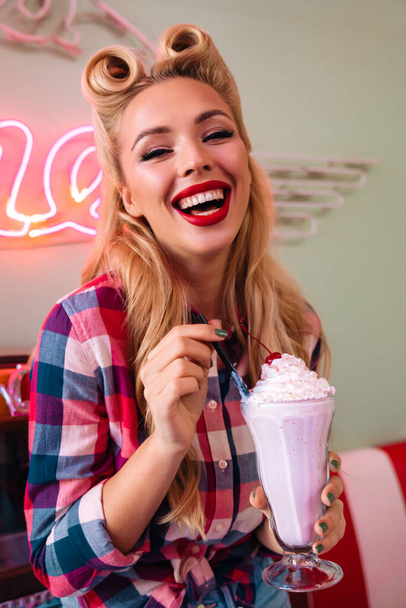 Photo of young cheerful woman with beautiful hairstyle drinking milkshake while leaning jukebox in retro american cafe - Фото, изображение