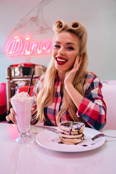Photo of gorgeous cheerful woman with beautiful hairstyle eating pancakes and drinking milkshake in retro american cafe - Fotografie, Obrázek