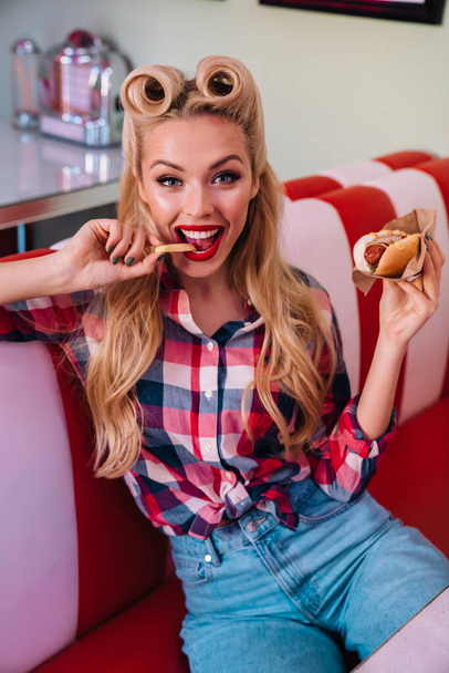Photo of charming joyful woman with beautiful hairstyle eating hotdog and french fries in retro american cafe - Photo, image