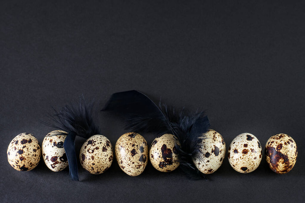 Huevos de codorniz con plumas negras sobre el fondo negro. Modo oscuro, textura minimalista negra, decoración de arte
. - Foto, Imagen
