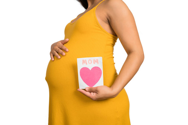 Close-up van een zwangere vrouw met een wenskaart tegen een witte achtergrond. Zwangerschap, nieuwe moeder en moederschap concept. - Foto, afbeelding