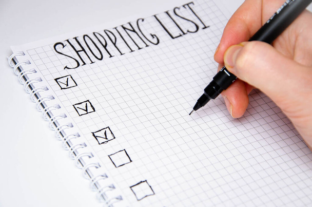 Shopping list. Hand holds a pen. Squared notebook with black pen on a white background. Record ideas, notes, plans, tasks. The list includes bread, milk, bananas. - Photo, Image
