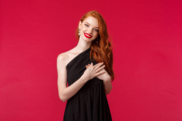 Fashion, luxury and beauty concept. Elegant good-looking redhead woman in black dress, with red lipstick and luxurious outfit, tilt head and smiling thankful, hold hand on heart touched with praises - Foto, Bild