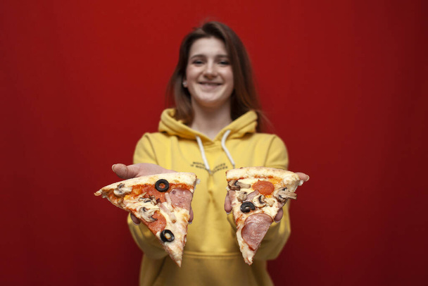 若い美少女は赤い背景においしいピザを食べ、スライスを与え、女性はファーストフード、クローズアップを食べることを提供しています - 写真・画像