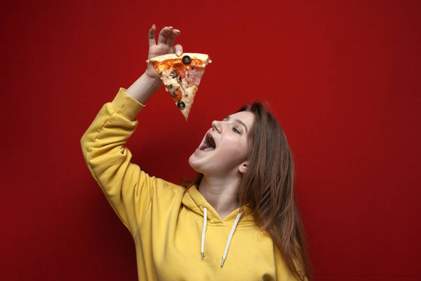 Young happy girl eats a piece of delicious pizza on a colored background, puts fast food in her open mouth - Fotó, kép