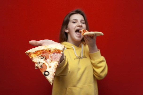 joven hermosa chica come sabrosa pizza sobre un fondo rojo y da una rebanada, una mujer se ofrece a comer comida rápida, primer plano
 - Foto, imagen