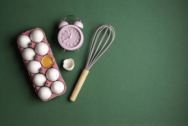 Time for cooking, flat lay, with pink alarm clock, whisk and an egg box. Fresh eggs on a green table. Home baking concept. - Photo, Image